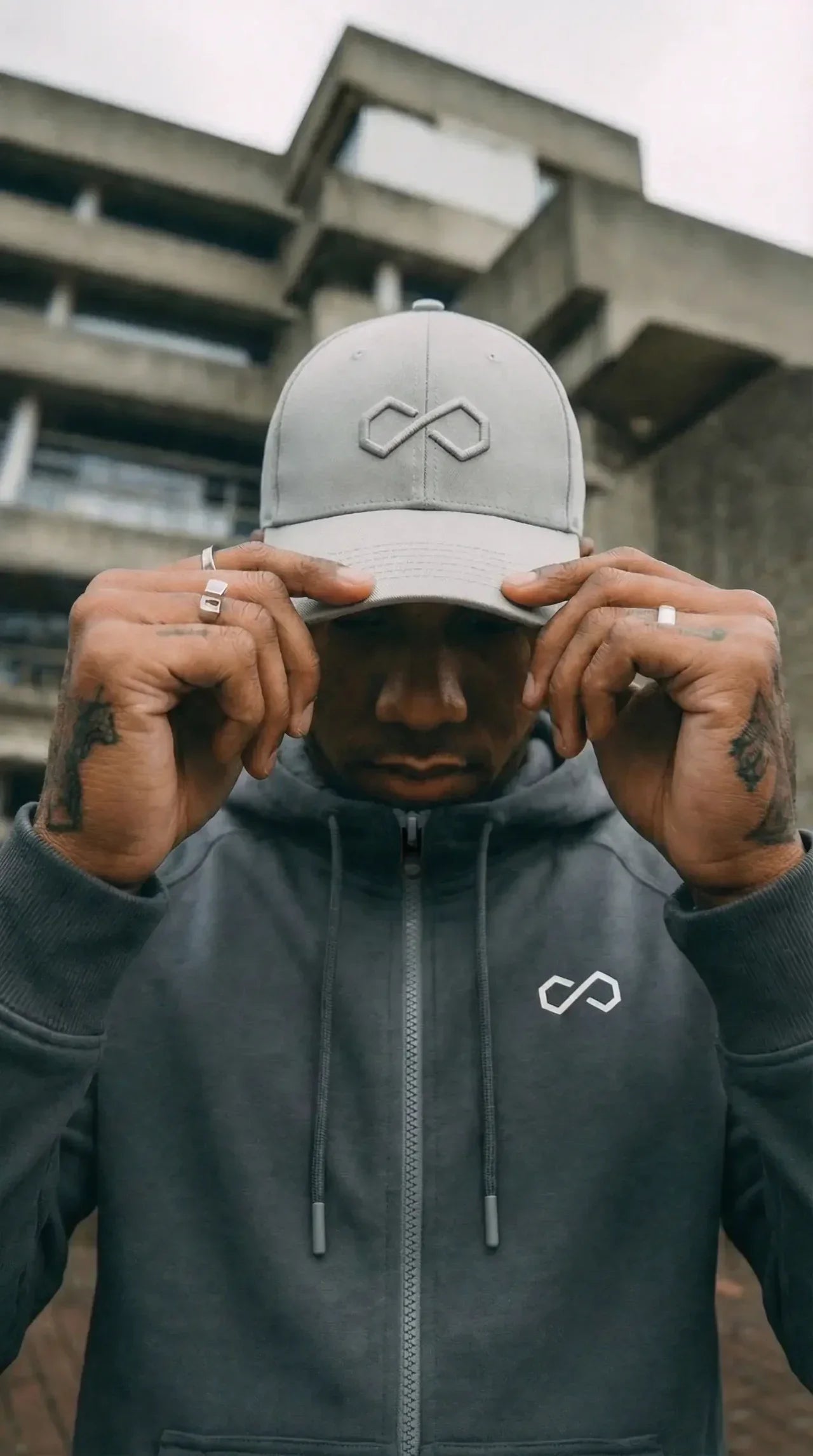 Homme portant une casquette grise et un hoodie gris foncé avec logo infini, style urbain