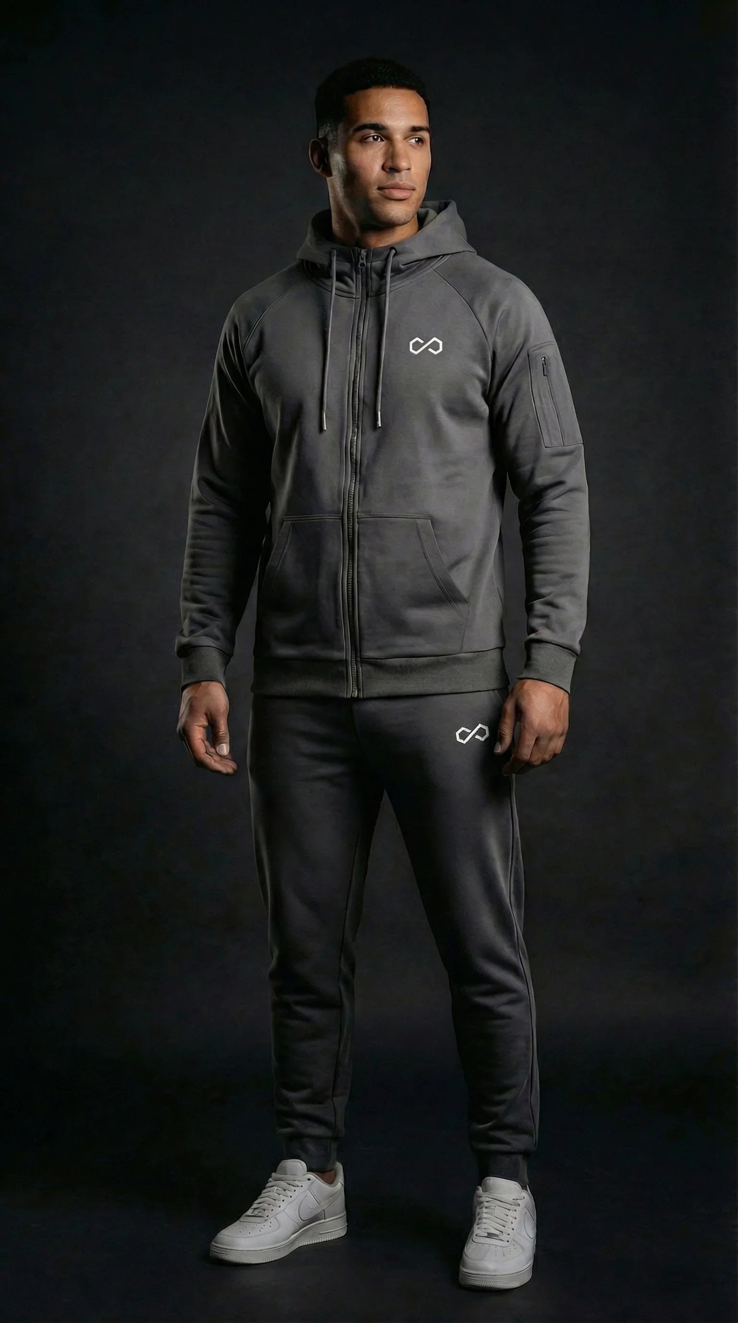Homme portant un ensemble de survêtement gris foncé avec capuche, logo minimaliste, sur fond noir.