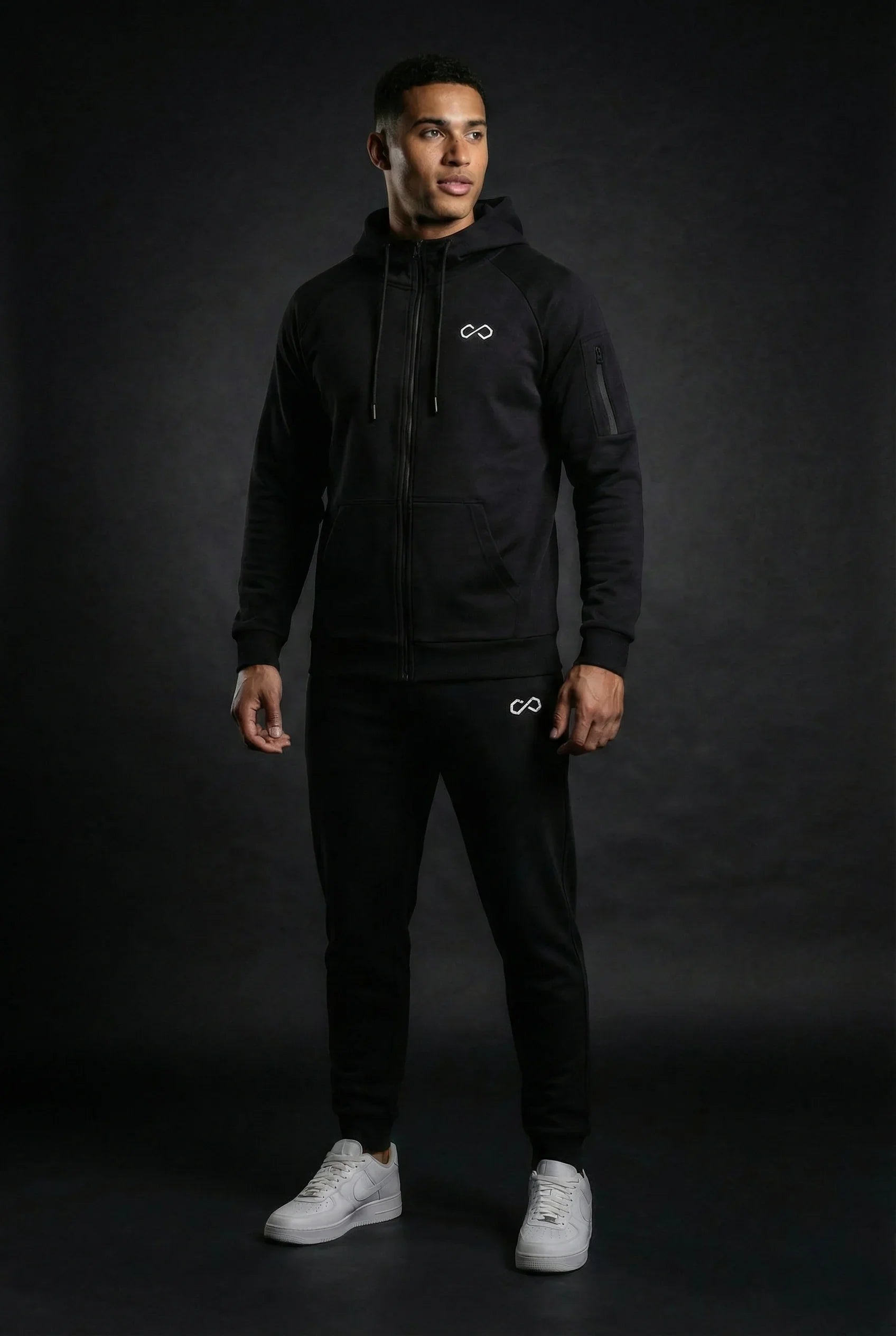 Homme portant un ensemble de survêtement noir minimaliste avec logo infini, fond sombre.