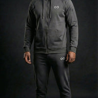 Homme portant un ensemble de survêtement gris foncé avec capuche, logo minimaliste, sur fond noir.